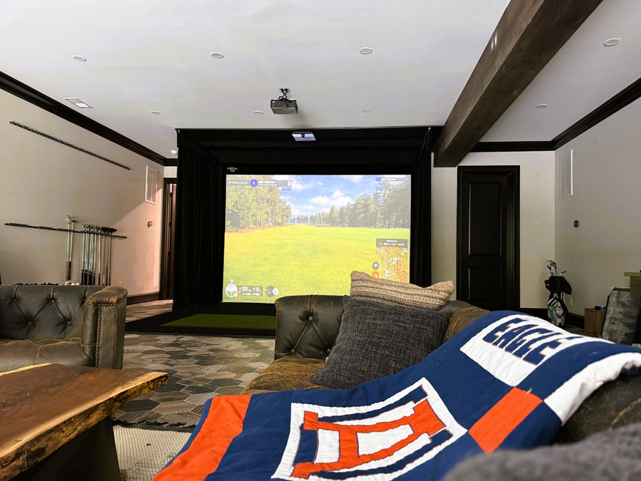 Golf simulator setup with a screen displaying a game, surrounded by comfortable furniture and a sports-themed blanket.