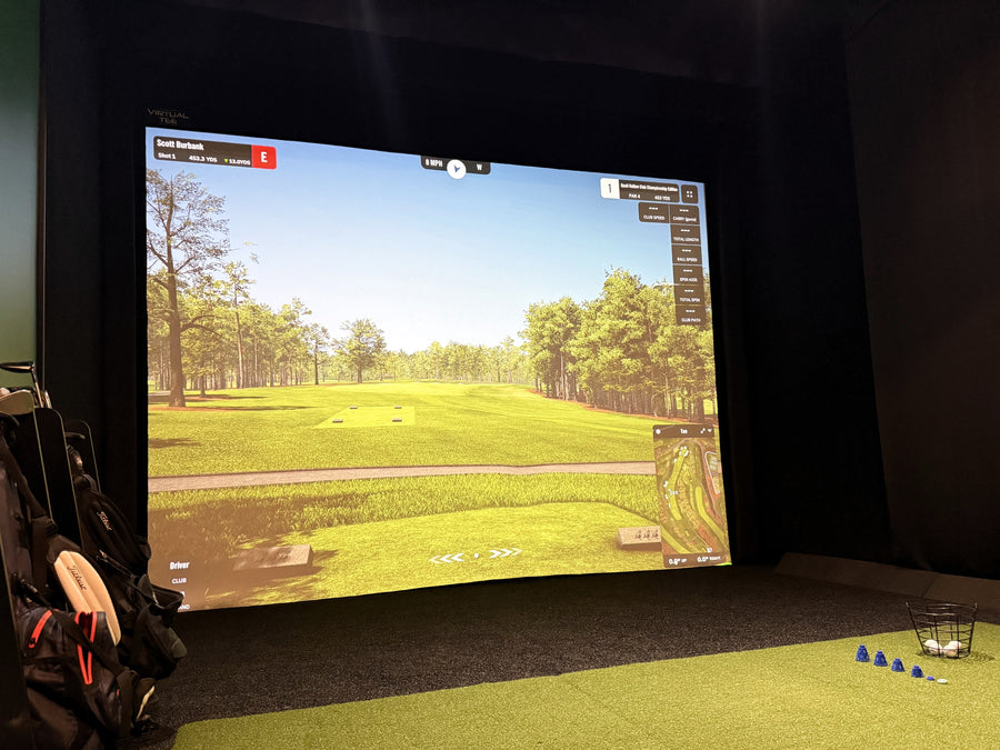 Golf simulator setup with screen displaying a golf course and golf clubs on the ground.