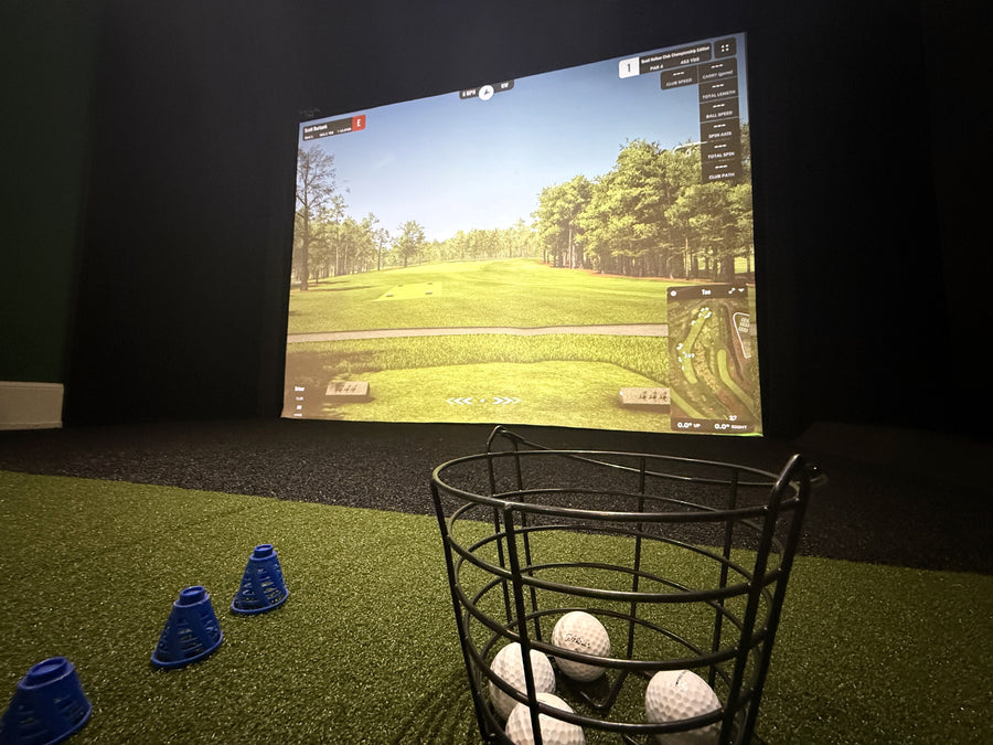 Golf simulator setup with screen displaying a golf course, balls, and cones on a green floor.