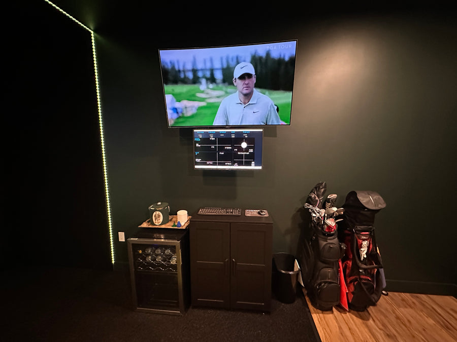 Golf simulator setup with two screens, golf bags, and audio equipment on a dark wall.