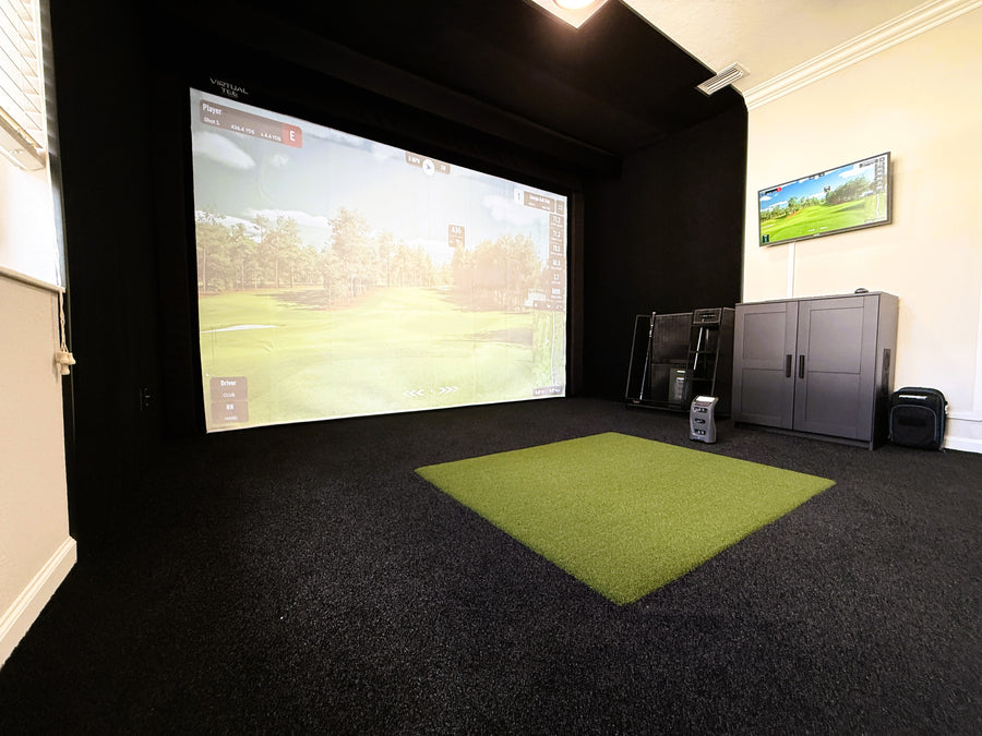 Indoor golf simulator setup with screen displaying a golf course and turf mat on the floor.