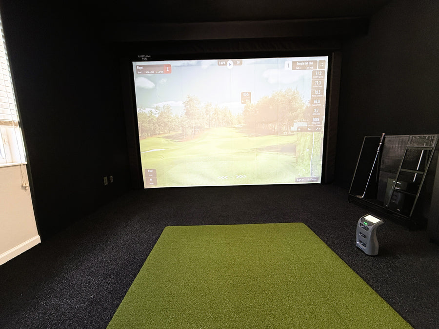 Indoor golf simulator setup with screen displaying a golf course and green mat on the floor.