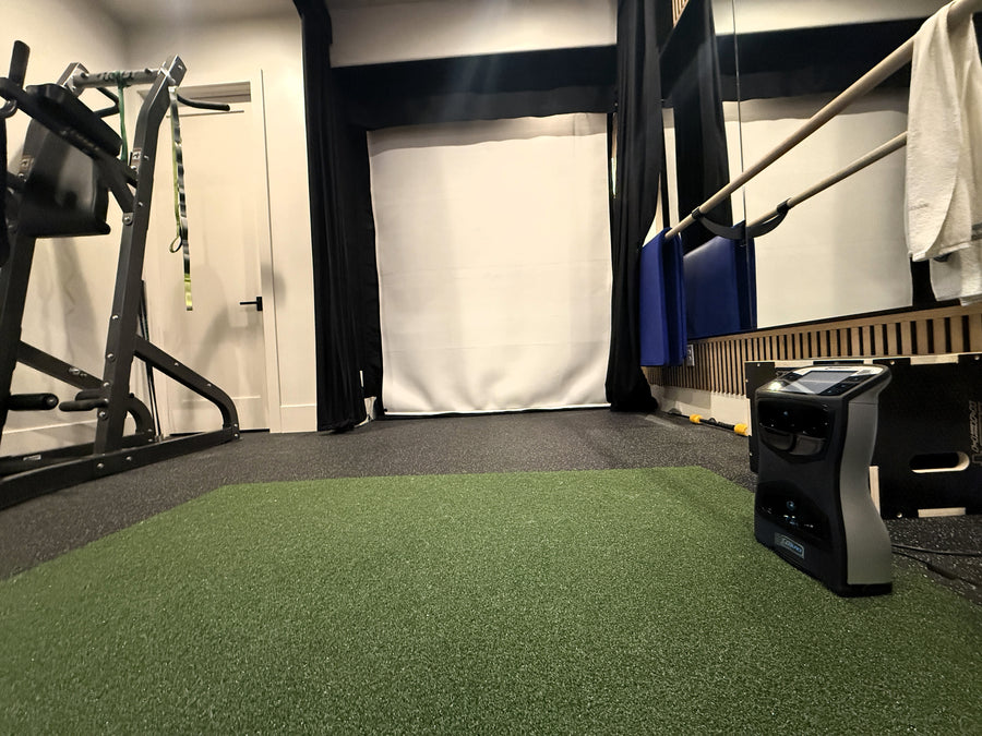 Gym golf simulator with green turf, exercise equipment, and white walls.