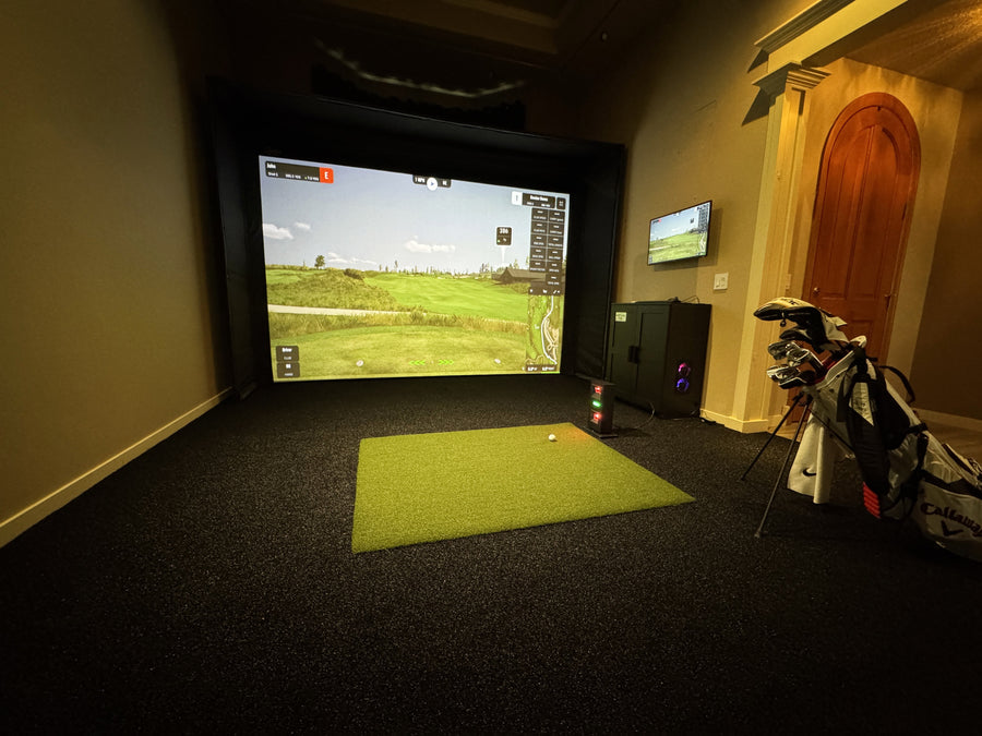 Indoor golf simulator setup with screen displaying a golf course, surrounded by golf bags and equipment.