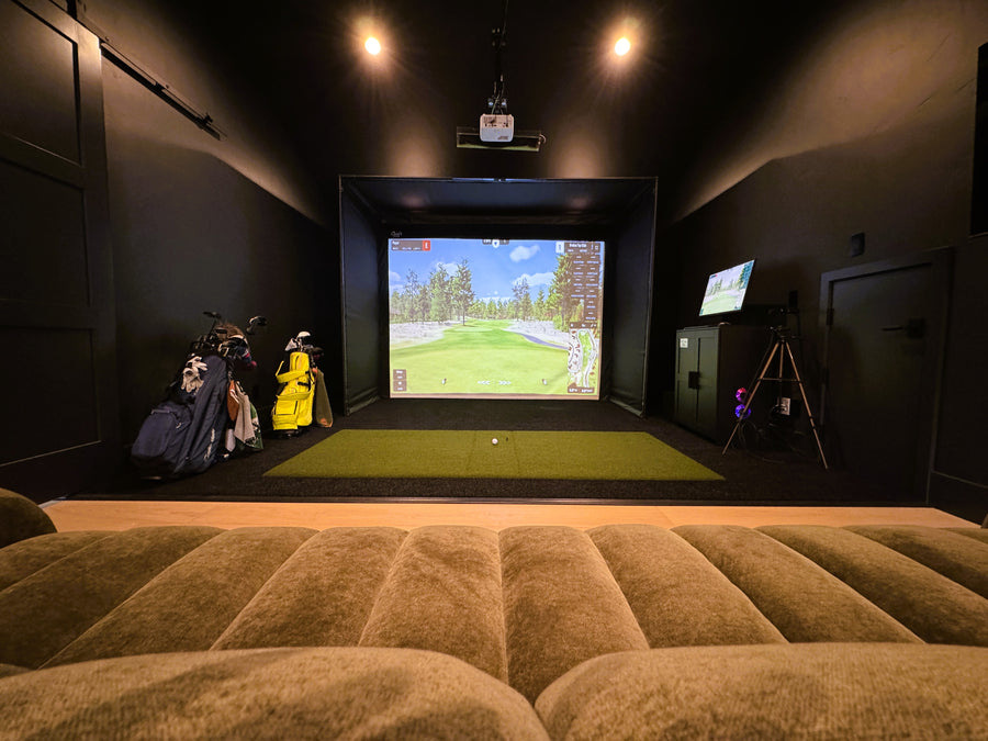 Indoor golf simulator setup with screen displaying a golf course, surrounded by golf equipment and a couch.