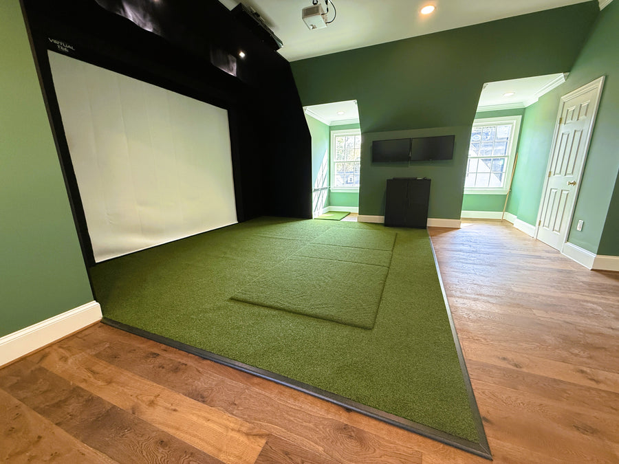 Indoor golf simulator setup with green turf and screen in a room with wooden flooring.