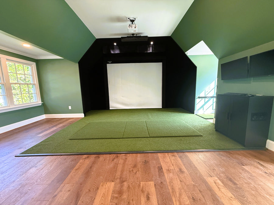 Indoor golf simulator setup in a room with green turf and wooden flooring.