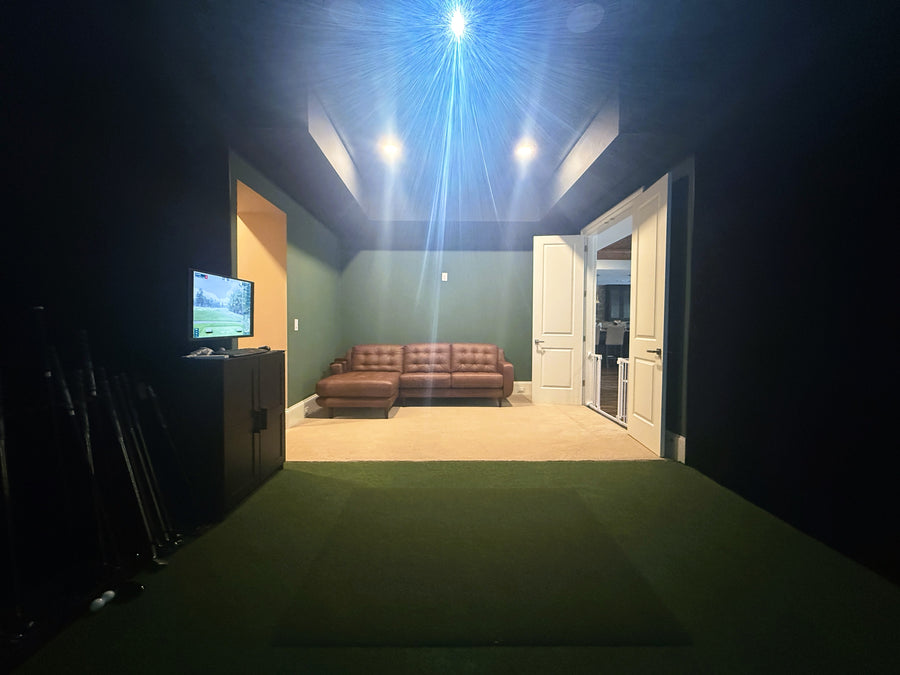 Indoor golf simulator with a brown leather sofa and TV, dimly lit.