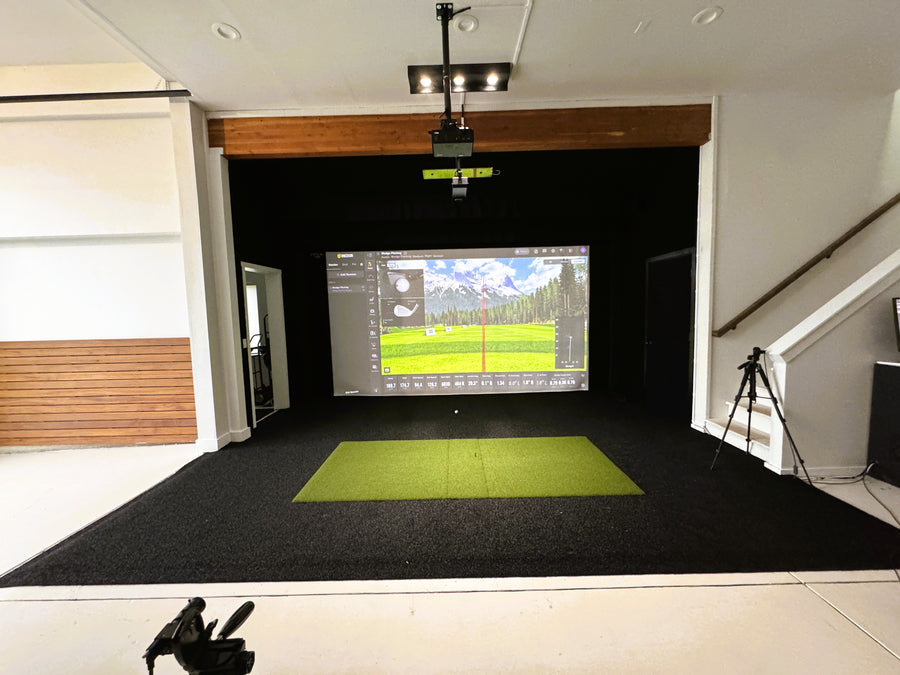 Indoor golf simulator setup with screen displaying a golf course on a black floor.