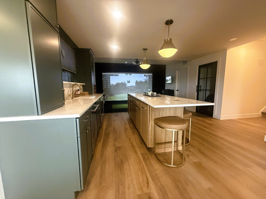 Modern kitchen with wooden flooring and a large island, golf simulator display in background.