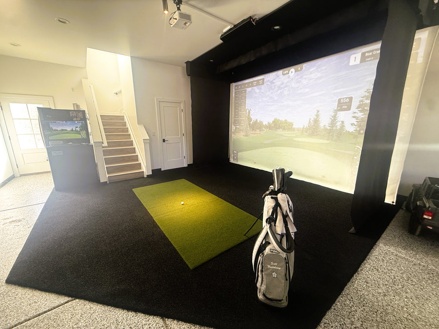 An indoor golf simulator setup with a large projected golf course screen, a green turf hitting mat, black carpeted floor, and a golf bag standing near the front right.