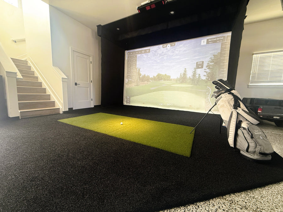 An indoor golf simulator setup with a bright green turf hitting mat, golf bag, and projector screen showing a golf course, in a home interior with stairs and a window.