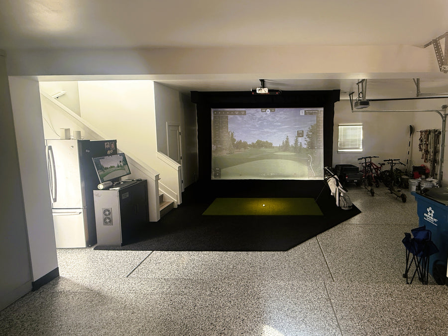 An indoor golf simulator setup in a home garage or basement, featuring a large projection screen showing a golf course, a black hitting box, a green turf mat, and a control console on the left. A refrigerator, staircase, and bicycles are visible in the surrounding garage space.