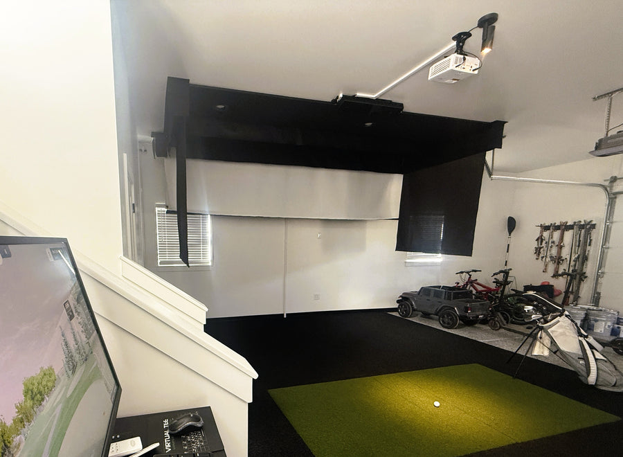 An indoor golf simulator setup with a large black projection screen mounted on a white wall, a projector overhead, a green turf golf simulator mat on the floor, and a golf cart with bicycles and equipment in the background.