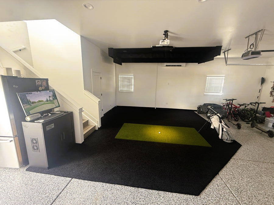 An indoor golf simulator setup in a garage, featuring a black overhead projector mounted on the ceiling, a projector on a stand with a TV screen showing a golf course, a black floor mat with a bright green turf target, a staircase on the left, and bicycles on the right.