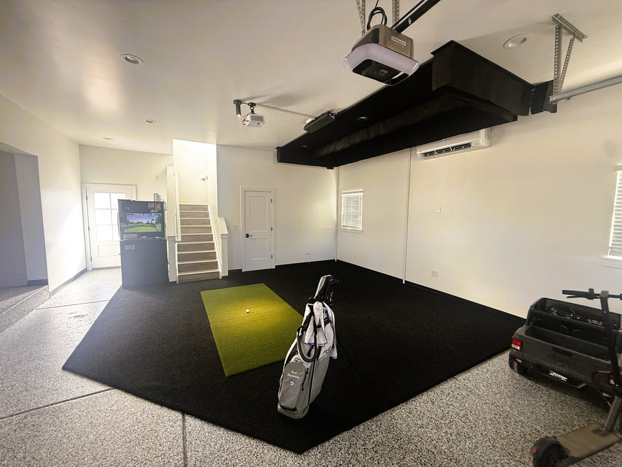 An indoor home golf simulator setup in a garage or basement, featuring a black turf mat with a green hitting box, a golf bag, a projector mounted on the ceiling, and a small vehicle in the corner.