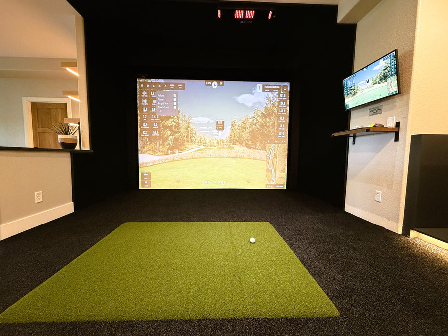 Indoor golf simulator setup with green turf and two screens displaying a golf course.