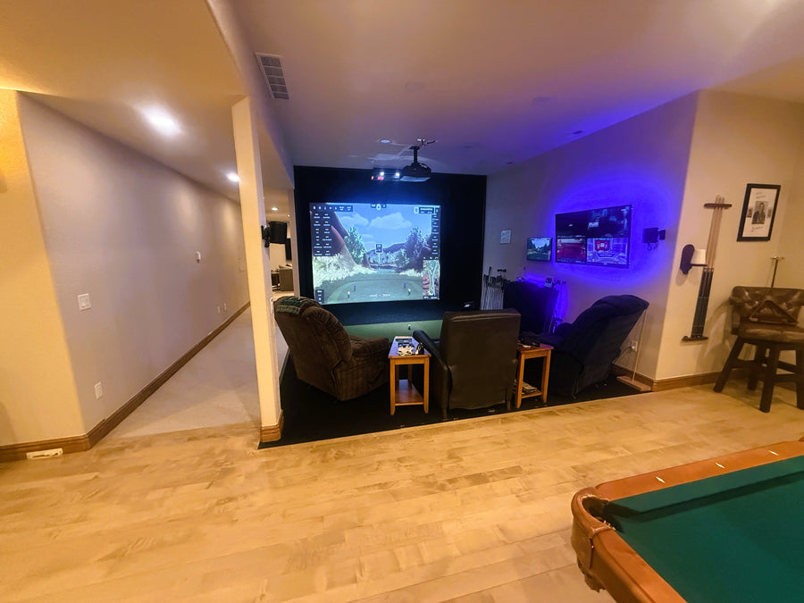 Indoor golf simulator with a large screen displaying a golf game, surrounded by comfortable seating and a pool table.