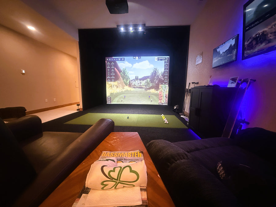 Golf simulator setup with a large screen displaying a golf round, surrounded by comfortable seating and decorative elements.