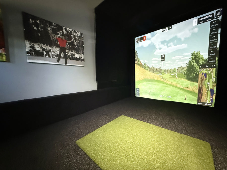 Indoor golf simulator setup with a screen displaying a golf course and a green mat on the floor.