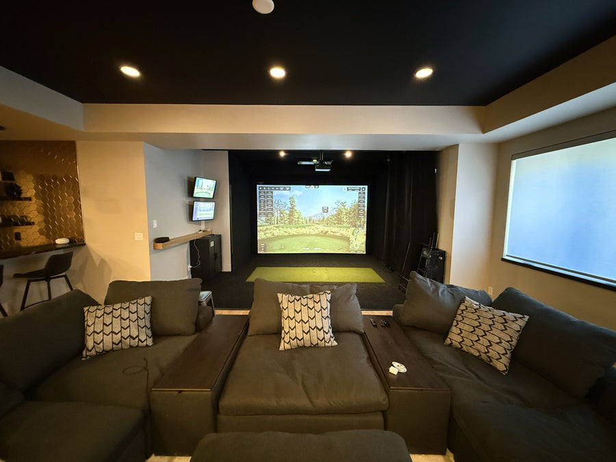 Modern golf simulator with a large screen displaying a video game, surrounded by comfortable seating and decorative elements.