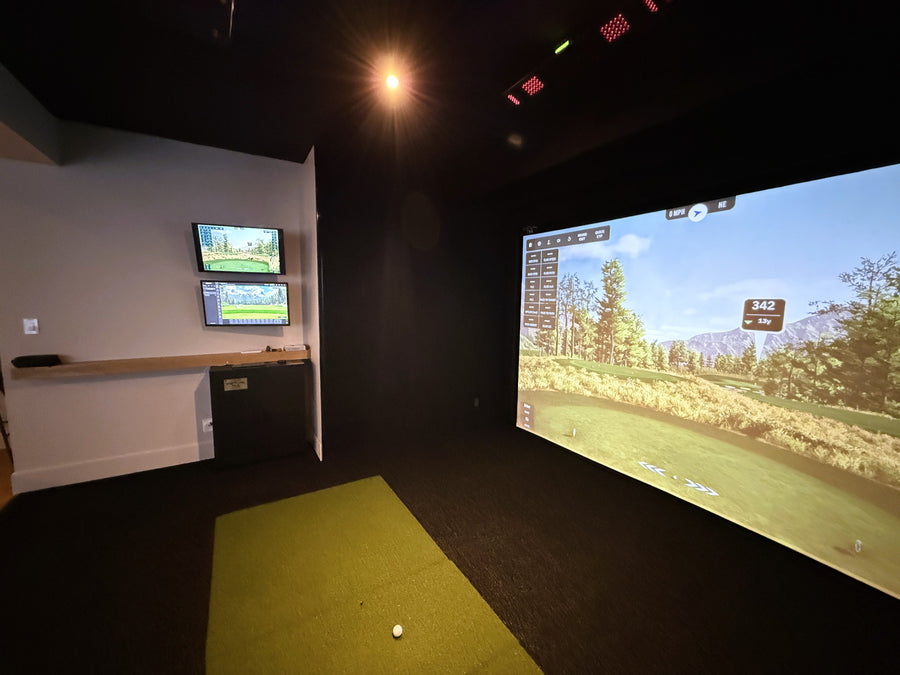 Golf simulator room with large screen displaying a golf course and two smaller screens on the wall.