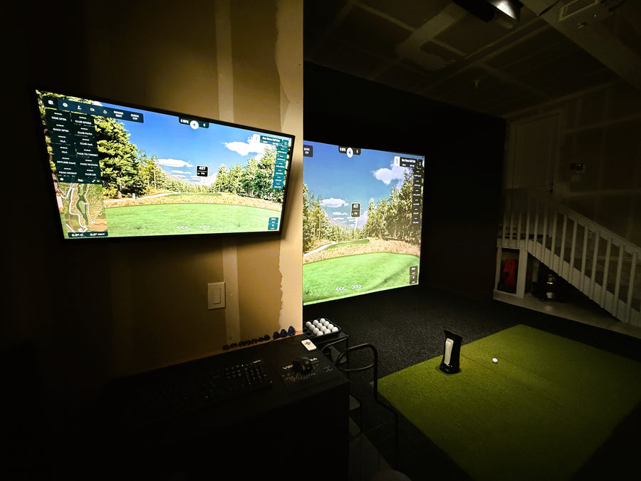 Golf simulator setup with two screens displaying a golf course in a dark room.