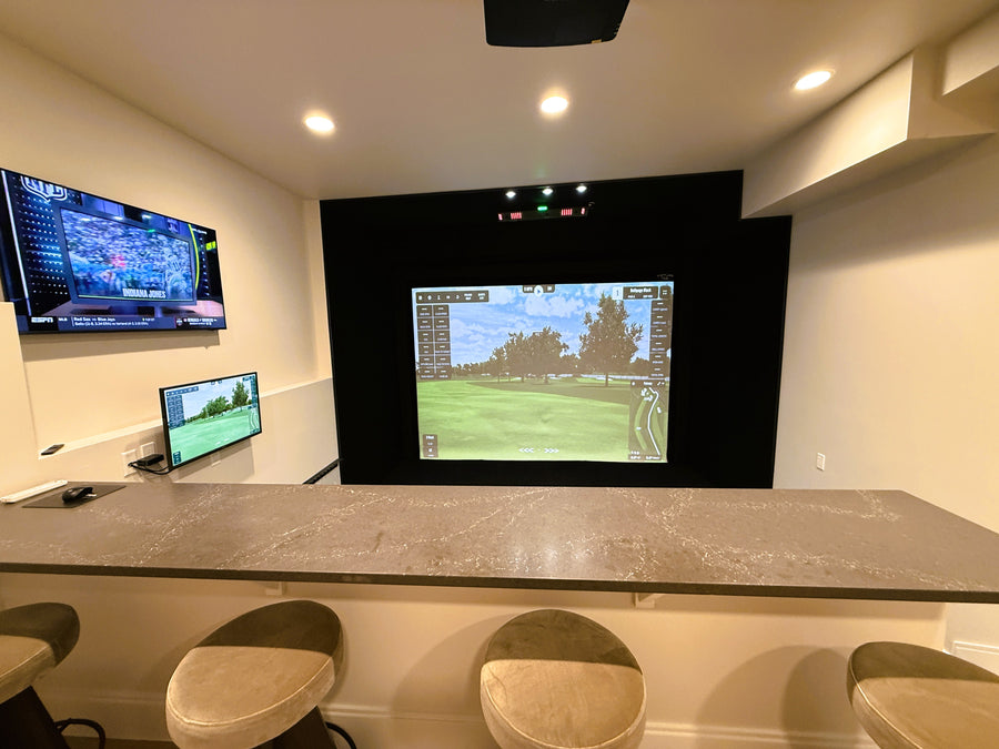 Golf simulator setup with multiple screens and a bar area in a room.