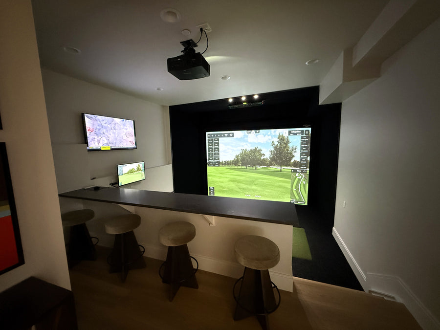 Golf simulator setup in a room with multiple screens and bar stools.