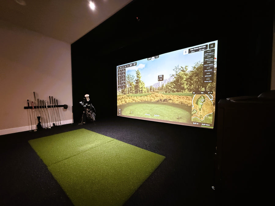Indoor golf simulator setup with screen displaying a golf course and golf clubs on a green mat.