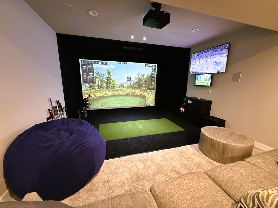 Golf simulator setup with a large screen displaying a golf game, surrounded by comfortable seating and decor.