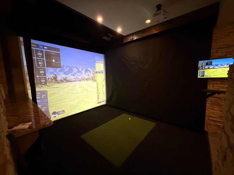 Indoor golf simulator setup with large screen and smaller monitor on wall.