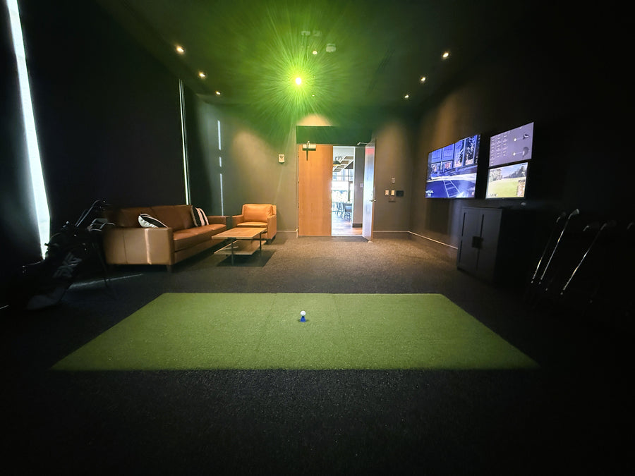 Indoor golf simulator with two televisions displaying sports content in a darkened room.
