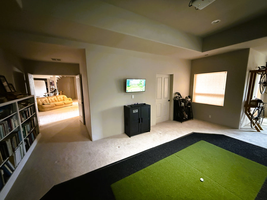 Indoor golf simulator with a green indoor putting green, TV, and bookshelf.