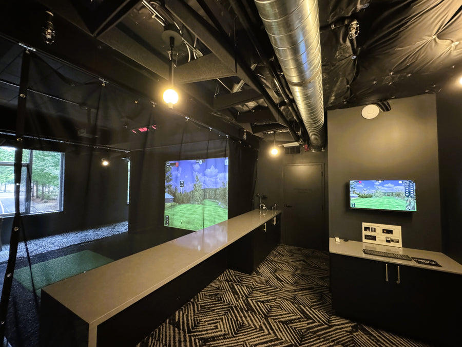 Indoor golf simulator room with multiple screens and a bench.