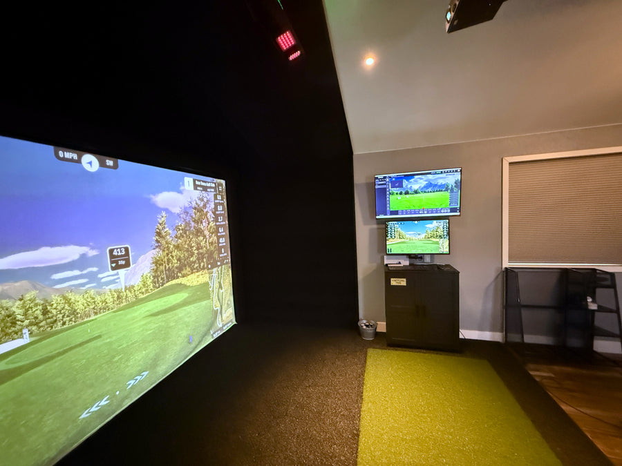 Indoor golf simulator setup with large screen and smaller monitor displaying game interface.