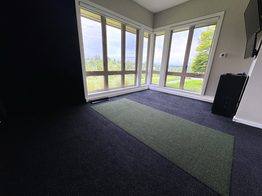 Corner of golf simulator room with Tv's mounted to the wall and projector to the ceiling. Green golf hitting mat on floor. 