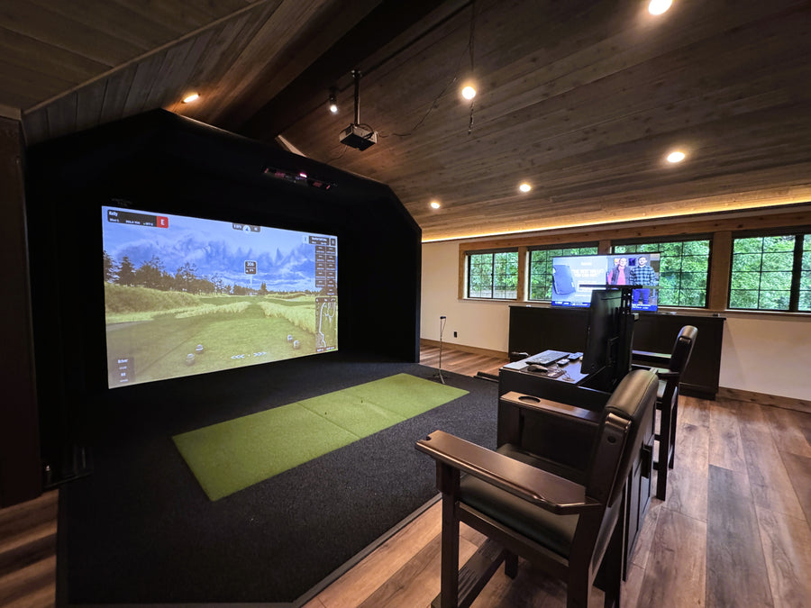 Golf simulator room with large screen displaying a golf simulation game, surrounded by chairs and turf.