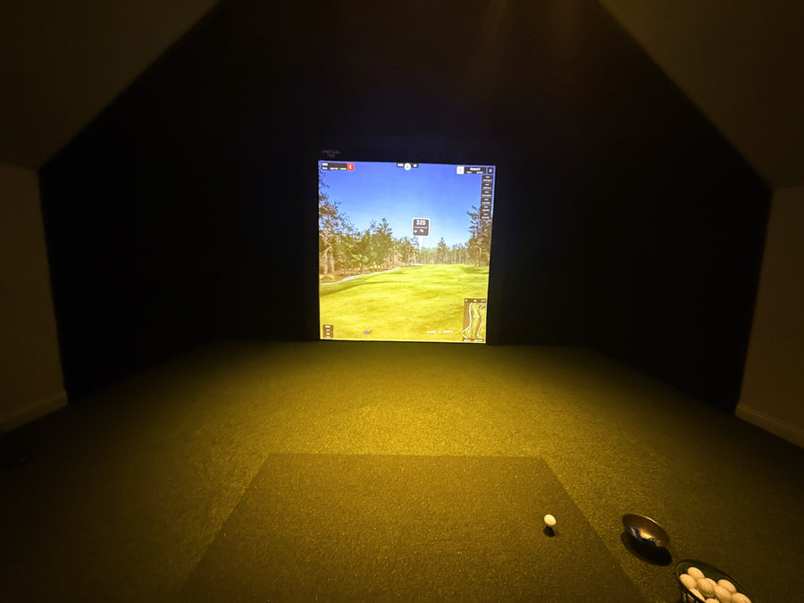 Golf simulator setup in a dark room with a screen displaying a golf course.