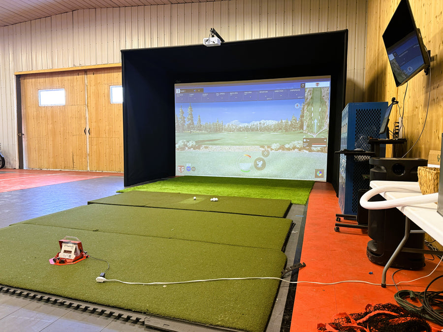 Golf simulator setup with screen, green mat, and equipment in a room.