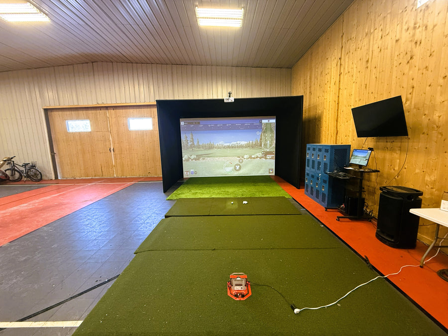 Indoor golf simulator setup with screen, turf, and equipment in a room with wooden walls and a red floor.