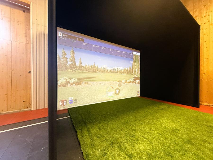 Indoor golf simulator with green turf and large screen displaying a golf course.