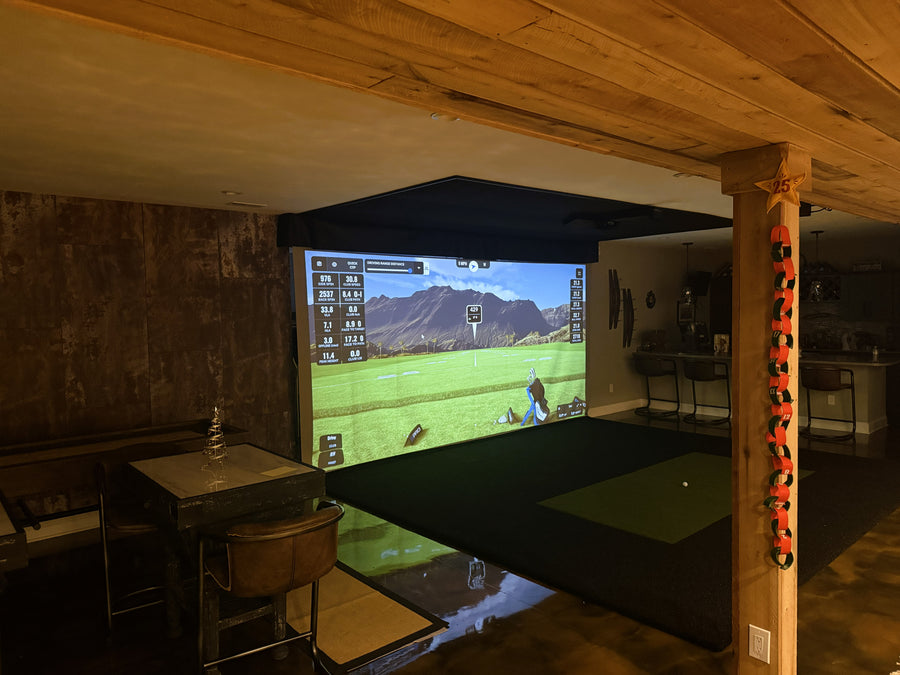 Indoor golf simulator setup in a room with wooden beams and a screen displaying a golf course.