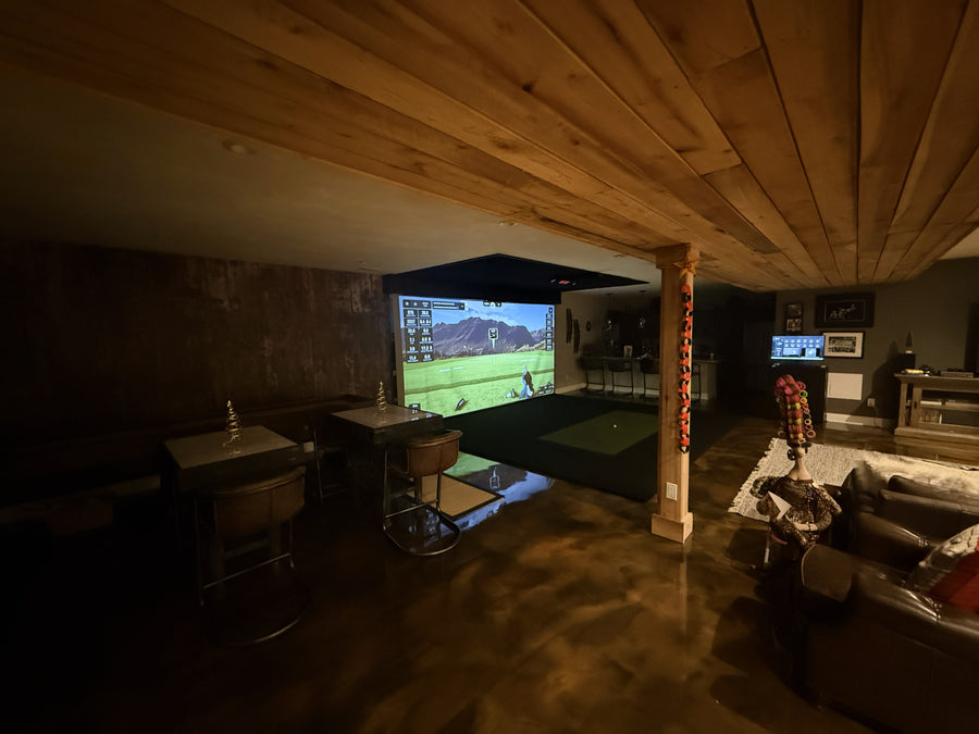 Indoor golf simulator setup with a large screen displaying a golf game, surrounded by furniture.