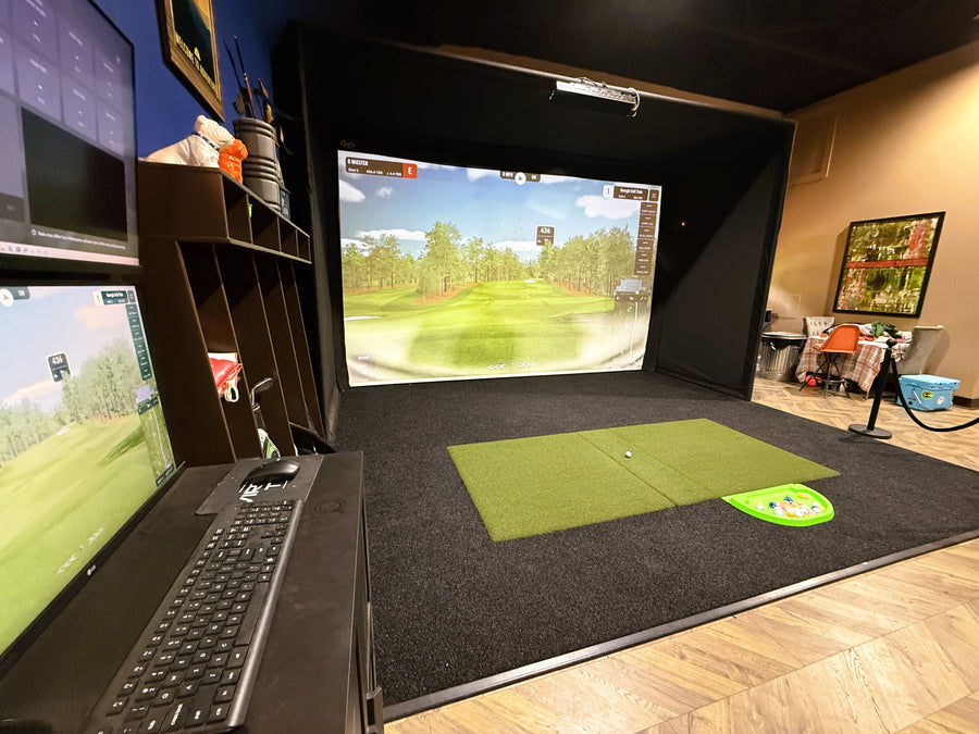 Golf simulator setup with two screens, a keyboard, and a mat on a wooden floor.