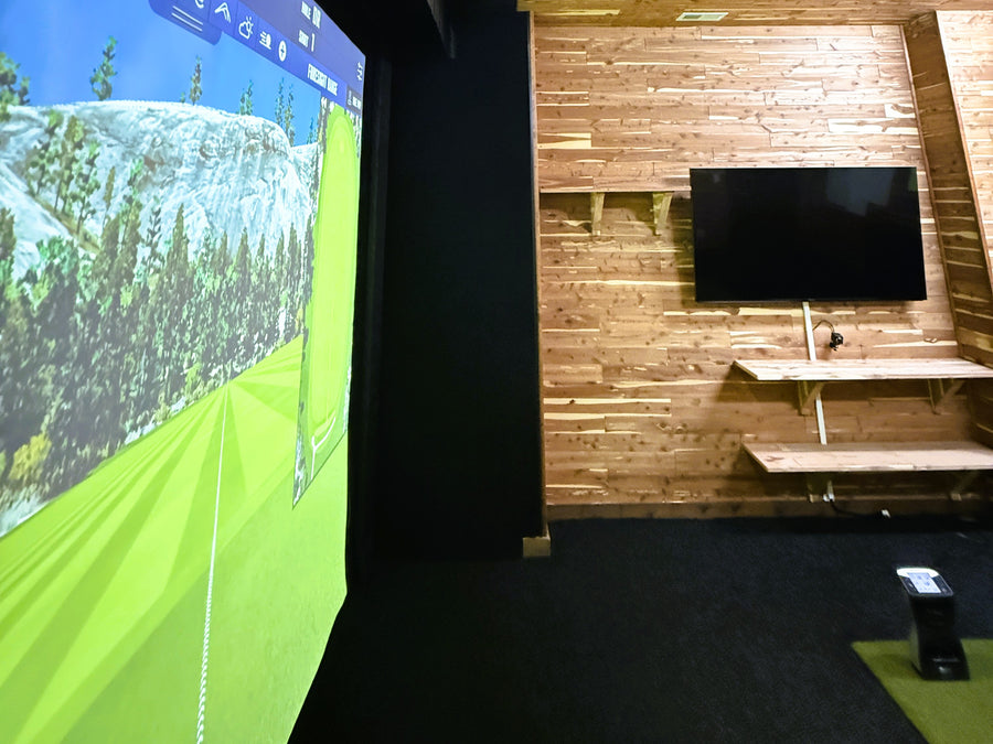 Golf simulator setup with screen displaying a golf course and TV on wooden wall.