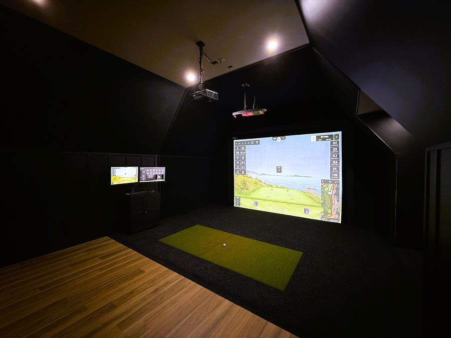Indoor golf simulator setup with projection screen and multiple monitors in a dark room.