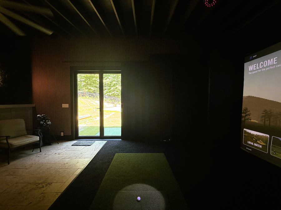 Indoor golf simulator room with screen displaying golf game to the right and seating to the left.