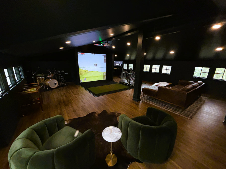 Golf simulator room with a large screen displaying a golf simulation game, surrounded by green chairs and wooden flooring.