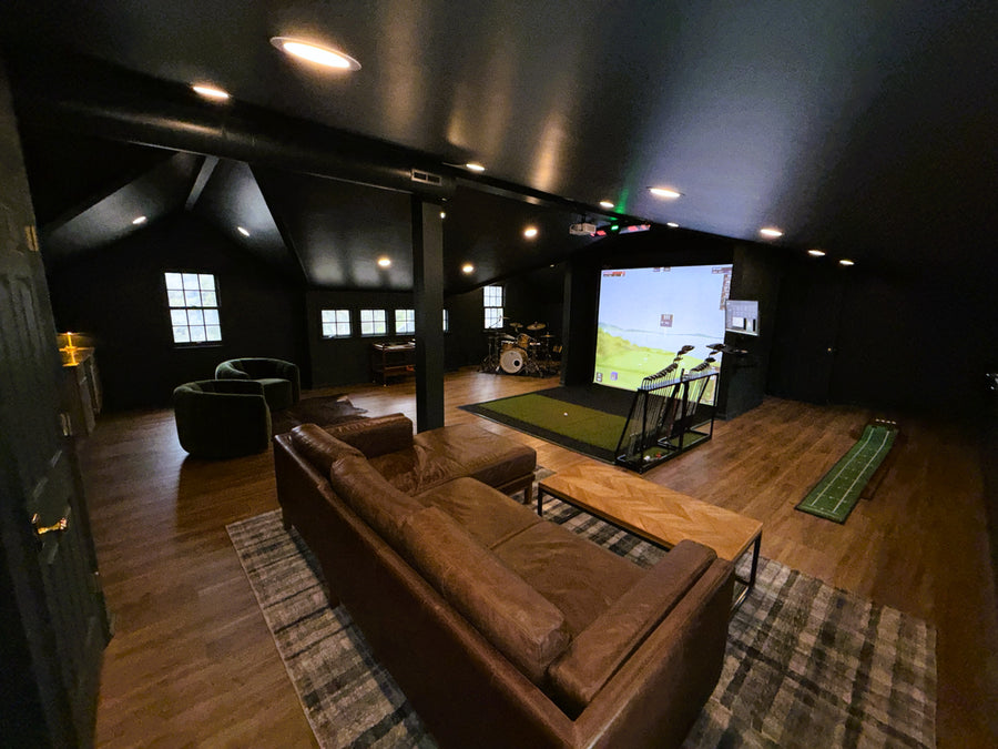 Golf simulator room with a large screen displaying a golf simulation game, brown sofa, and sports ball.
